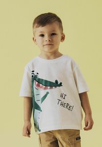 Camiseta blanca con un gráfico de un caimán de dibujos animados en verde y el texto "¡HOLA!" en negro. El niño lleva pantalones cortos caqui y está de pie frente a un fondo amarillo.