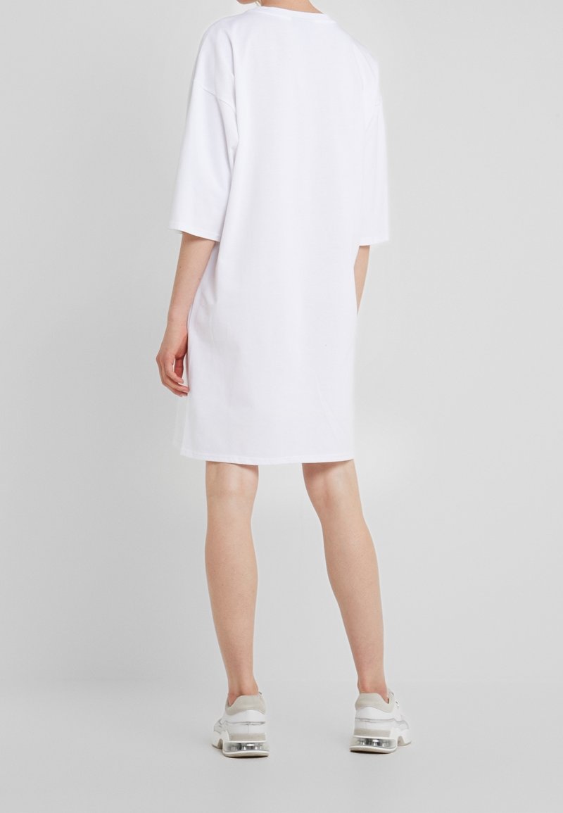 Osoba w prostym, białym, oversize'owym sukience typu T-shirt i białych sneakersach, stojąca tyłem na jednolitym szarym tle.