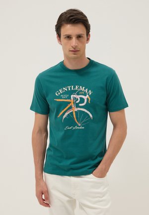 Homme portant un t-shirt sarcelle avec un graphisme de vélo orange et le texte "Gentleman Bicycle Race 1976 East London" et un pantalon blanc, debout devant un fond uni.