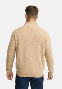 Pull en tricot beige avec un col montant, texture côtelée, manches longues et coupe décontractée, porté sur un jean bleu foncé.