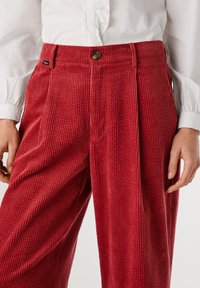 Sarkanas korduroja bikses ar vertikālu ribošanu, priekšējām krokām un sānu kabatām, akcentētas ar brūnu pogu un mazu zīmola birku netālu no jostas.