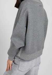 Sudadera gris con un corte relajado, que presenta un cinturón y puños de canalé. Sutiles acentos brillantes en el dobladillo. Tejido suave y texturizado.