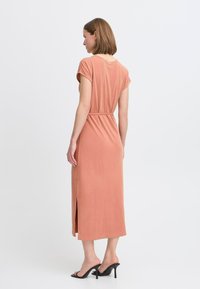 Perzikkleurige midi-jurk met korte mouwen, een ceintuur in de taille, een zijsplit en zachte stof. Gerd gedragen met zwarte hoge hakken.