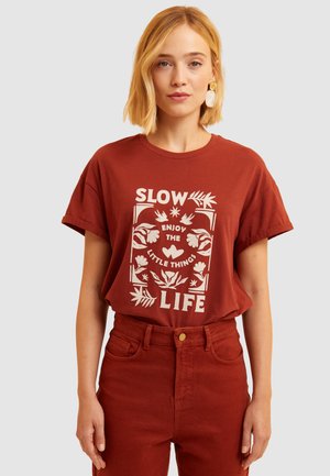 T-shirt de couleur rouille à manches courtes avec du texte blanc et des graphismes floraux, associé à un pantalon taille haute assorti.