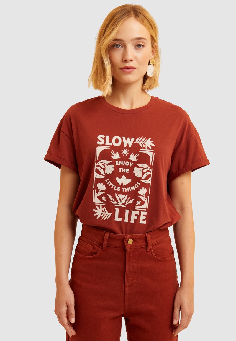Camiseta de color óxido con mangas cortas que presenta texto blanco y gráficos florales, combinada con pantalones de cintura alta a juego.