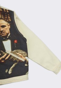Felpa nera e beige con una grafica prominente di un uomo che tiene in braccio un gatto grigio, presenta una zip su un lato e dettagli vivaci.