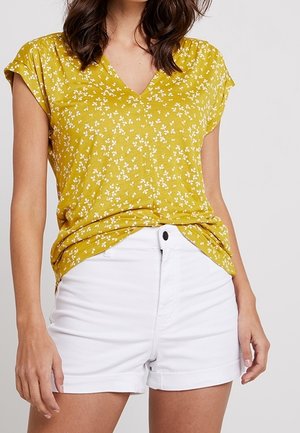 Frau trägt ein gelbes, kurzärmeliges V-Ausschnitt-Oberteil mit Blumenmuster, das in eine weiße, hoch taillierte Shorts mit Knopfverschluss gesteckt ist.