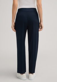 Marineblaue maßgeschneiderte Hosen mit glatter Textur, ausgestattet mit einem hohen Bund und zwei Gesäßtaschen. Getragen mit hellen Sneakers.