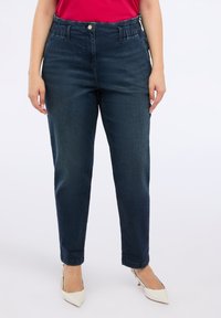 Dunkelblaue, taillierte Jeanshose mit hohem, elastischem Bund, geradem Bein und seitlichen Taschen. Kombiniert mit weißen High Heels.