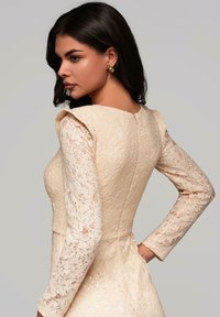 Beige spetsklänning med långa ärmar, en figursydd bodice och texturerad spetsdetaljering. Har en rundad halsringning och dragkedja bak.