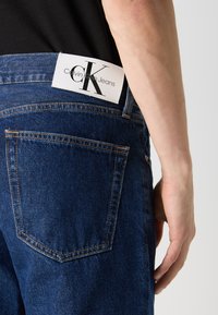 Tumedast sinisest denimpükstest, millel on valge silt "CK" vööl, aktsendiga oranži õmbluse ja ühe tagataskuga.