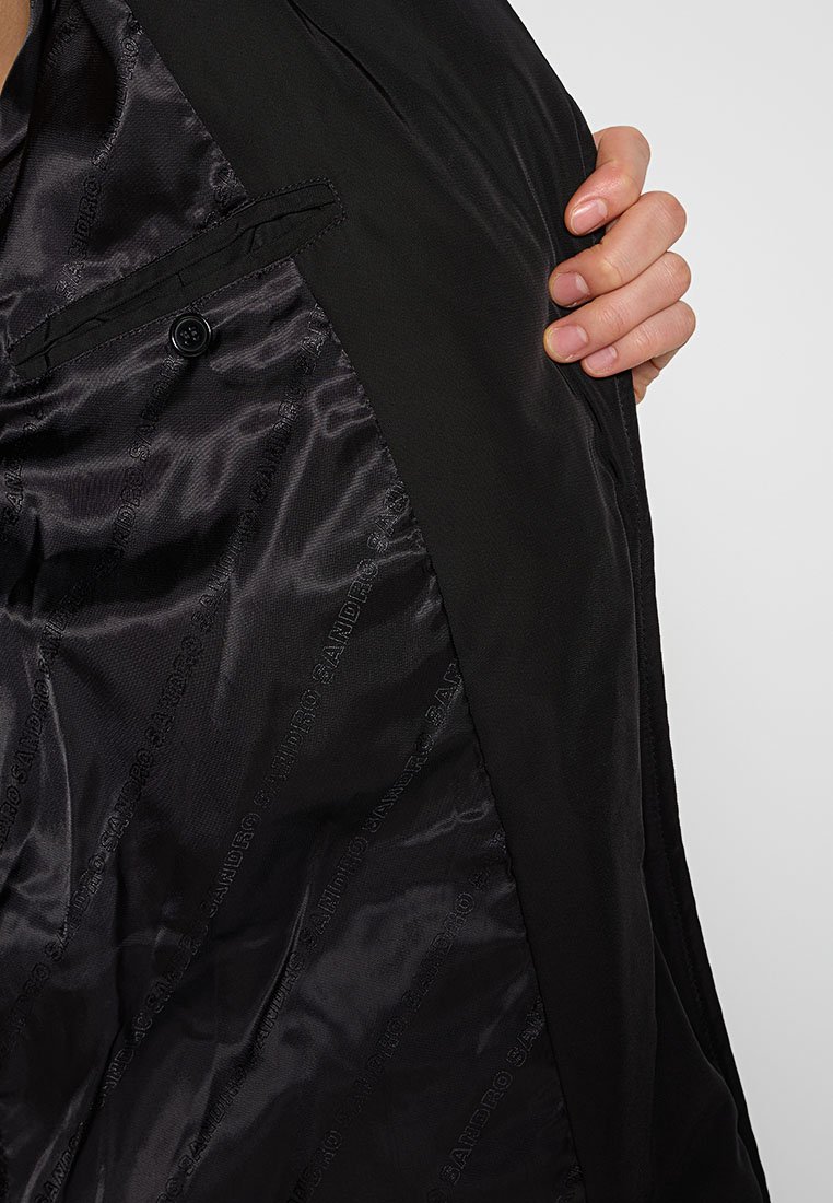 Schwarze Jacke mit einem glatten Satinfutter und einem dezenten Logomuster. Verfügt über eine Außentasche und einen Knopf auf der Brust.