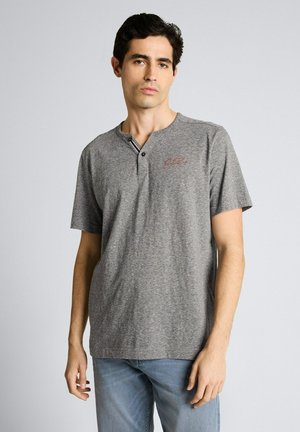 Młody mężczyzna ubrany w szarą koszulkę henley z krótkim rękawem i cienkimi paskami oraz jasnoniebieskie dżinsy, stojący na jednolitym tle.