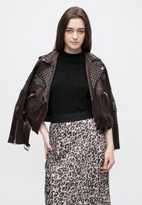 Młoda kobieta ubrana w brązową skórzaną kurtkę z ćwiekami, czarny top i spódnicę w panterkę, stoi na tle jednolitego białego tła.