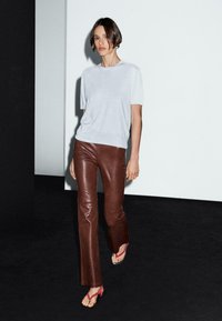 Una donna con capelli corti cammina con sicurezza indossando un top grigio chiaro e pantaloni di pelle marrone, su uno sfondo minimalista.