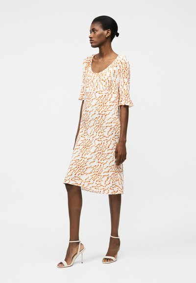 Γυναίκα που στέκεται, φορούσα λευκό φόρεμα μέχρι το γόνατο με πορτοκαλί λουλουδένιο σχέδιο, λαιμόκοψη σε σχήμα φραμπαλά, μανίκια με βολάν μέχρι τους αγκώνες και λευκά τακούνια με λουράκι στον αστράγαλο.