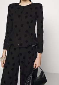 Černý sametový kabát s přiléhavým střihem, s předním zipem a jemnými vzory listů. Doplněný o černé kalhoty ve stejném stylu a černou kabelku.