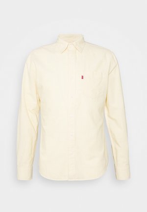 Chemise beige à manches longues avec boutons, col et poche poitrine dotée d'une petite étiquette rouge sur le côté gauche.
