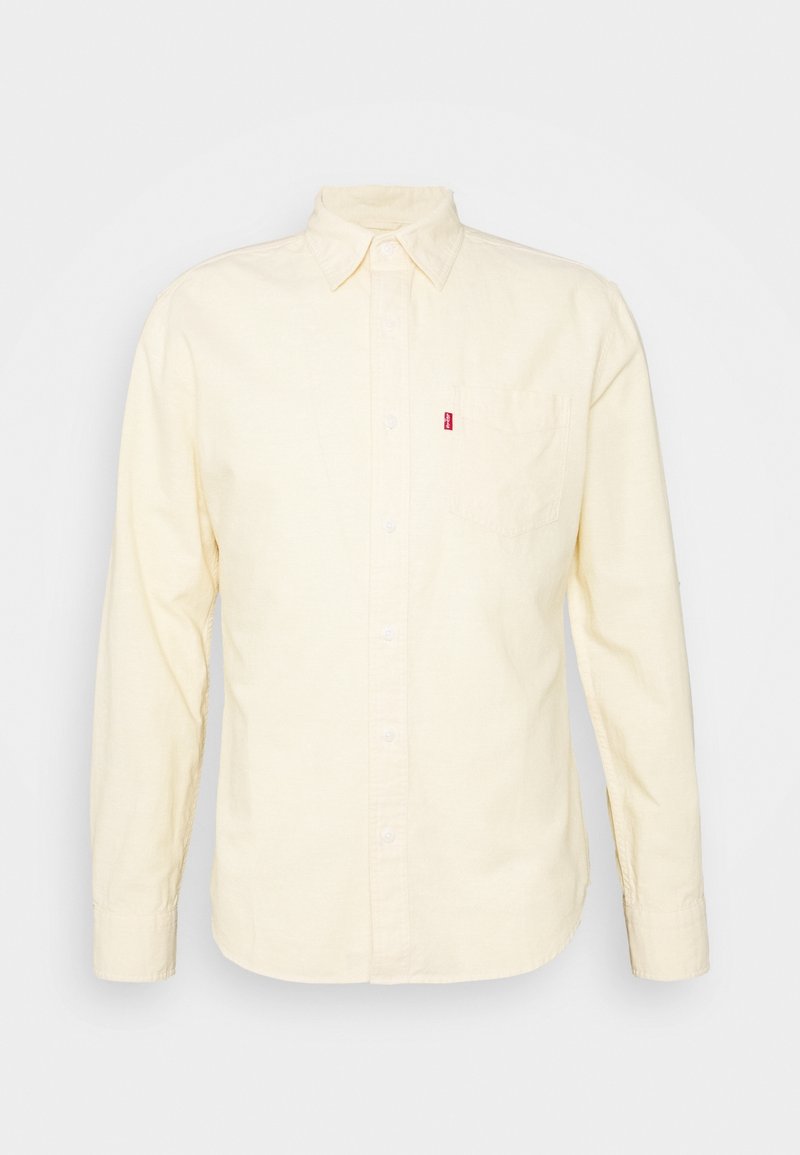 Chemise beige à manches longues avec boutons, col et poche poitrine dotée d'une petite étiquette rouge sur le côté gauche.