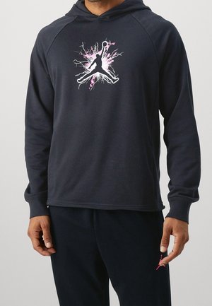 Mann trägt schwarzen Hoodie mit pink-weißem elektrischen Basketballspieler-Logo und passenden schwarzen Hosen mit kleinem pinkfarbenem Logo am Oberschenkel.