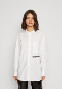 Biała koszula z guzikiem i kołnierzykiem, z kieszonką na piersi oraz czarnym logo "KARL LAGERFELD JEANS" z przodu. Gładki, wyrazisty materiał.