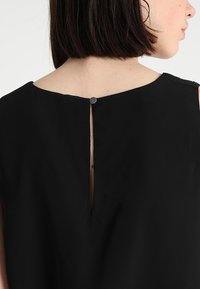 Svart ärmlös klänning med en enkel design bak med en vertikal slits och en rund knappstängning vid nacken. Mjukt material.