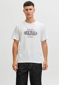 Hombre vistiendo una camiseta blanca con la inscripción "ORIGINALS" y unos vaqueros oscuros, de pie contra un fondo claro y liso.