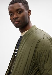 Giacca bomber trapuntata verde con colletto a coste, dotata di chiusura con zip. Indossata sopra una camicia a righe blu navy e bianche.