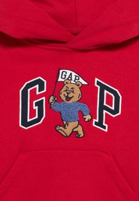 Κόκκινο φούτερ με κουκούλα με κεντημένη αρκούδα σε μπλε πουλόβερ που κρατάει μια σημαία που γράφει "GAP" ανάμεσα στα μεγάλα γράμματα "G" και "P".