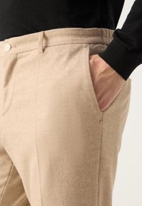 Béžové kalhoty na míru vyrobené z texturované látky, s bočními kapsami a zapínáním na knoflík. Pas je elastický pro pohodlí.