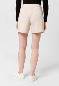 Béžové bavlněné kraťasy s elastickým pasem, mají volný střih a čistý lem, doplněné o bílé tenisky na jednobarevném pozadí.