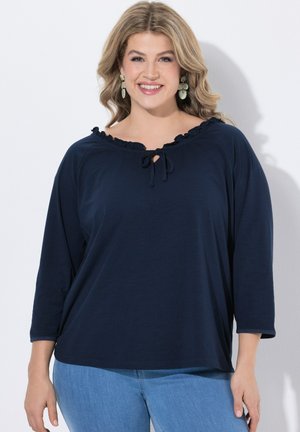 Mujer sonriente con cabello rubio ondulado que lleva una blusa azul marino oscura con cuello anudado y mangas 3/4 combinada con vaqueros azul claro.