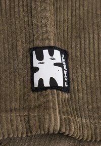 Καφέ υφασμάτινο κορδόνι από κοτλέ με ένα ασπρόμαυρο μπαλώματα που δείχνει αφηρημένα πρόσωπα και τη λέξη "FORMET" στην άκρη.