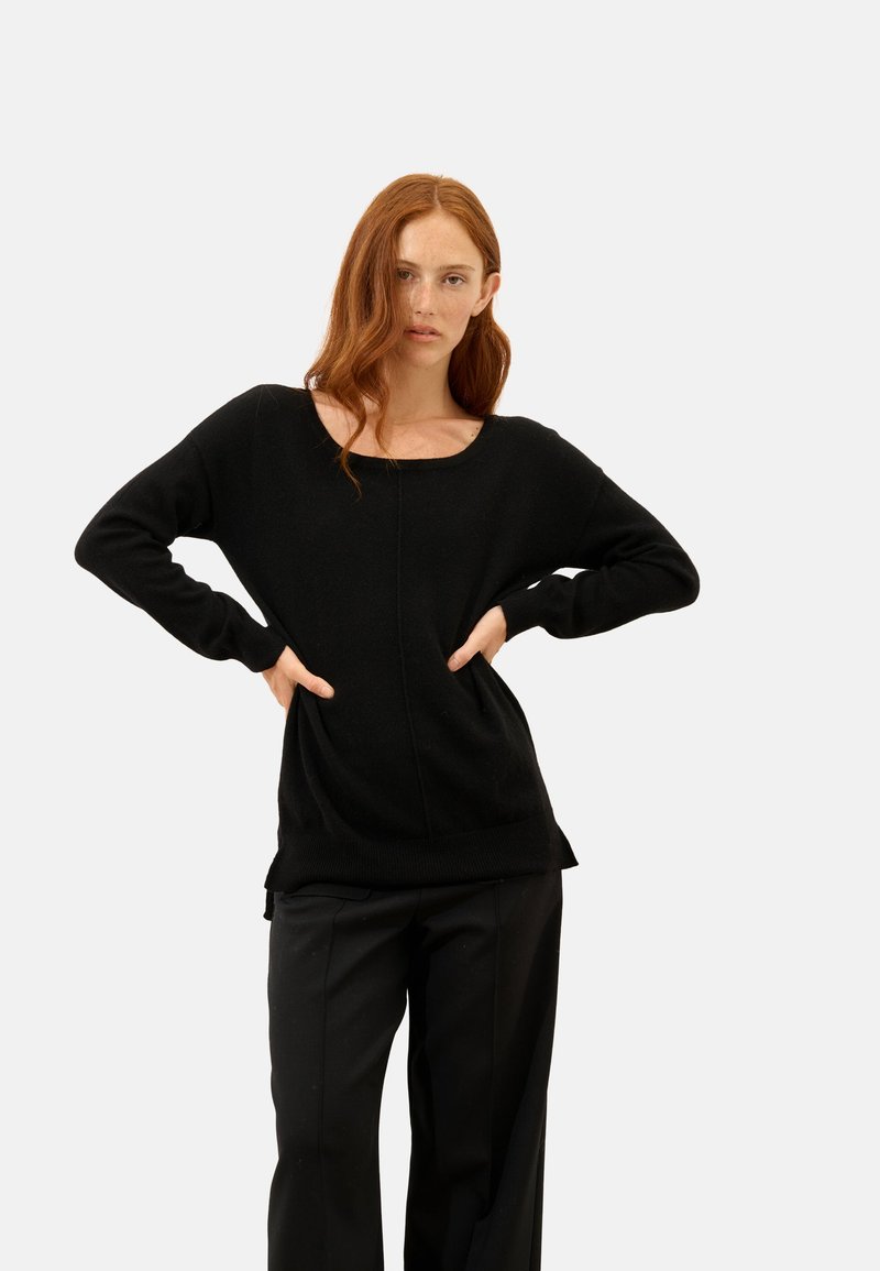 Schwarzer Strickpullover mit weitem Ausschnitt, gerippten Bündchen und entspannter Passform, kombiniert mit locker sitzenden schwarzen Hosen. Schlichte, glatte Textur.