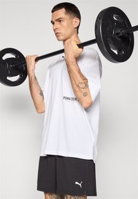 Hvit oversized t-skjorte med "PUMA CREW" logo, kombinert med mørkegrå shorts med hvit Puma-logo. Vektløfting med svart vektstang.