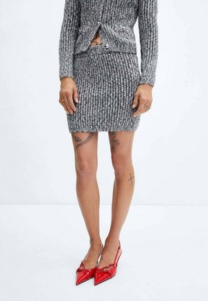 Personne portant une jupe en maille côtelée grise et un haut assorti, montrant des tatouages sur les jambes, talons slingback rouges pointus, debout sur fond blanc.