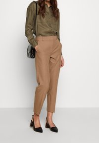 Žena v zkrácených camel kalhotách, olivově zelené košili na knoflíky, černých slingback podpatcích a nese malou černou kabelku přes rameno.