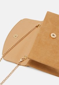 Pochette en daim camel avec une bandoulière chaîne en or. Dotée d'une fermeture à pression et d'un intérieur simple. Texture lisse avec un minimum de ferrures.
