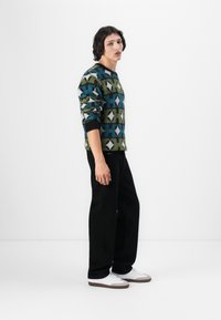 Strikkegenser med geometriske mønstre i grønt, blått og hvitt. Sorte bukser; hvite sneakers med grå detaljer. Modell stående sideveis.