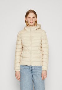 Lys beige dunjakke med hette, med glidelås foran, ribbestrikkede mansjetter og quiltet tekstur. Bært over blå denimjeans.