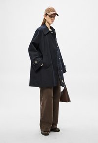 Ναυτικό παλτό κουμπωτό με μεγάλο γιακά, δύο μπροστινές τσέπες και χρυσαφένιες λεπτομέρειες στα κουμπιά, συνδυασμένο με καφέ παντελόνια φαρδιάς γραμμής και καφέ καπέλο.