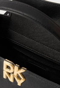 Μαύρη τσάντα χειρός από υφασμάτινη υφή με χρυσή ένδειξη λογότυπου. Το εσωτερικό περιλαμβάνει μια τσέπη με φερμουάρ και λεπτομέρειες λογότυπου.