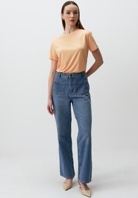 Brzoskwiniowa koszulka z krótkim rękawem w połączeniu z jeansami z wysokim stanem w niebieskim kolorze, z kieszeniami z przodu i z tyłu. Modelka nosi beżowe czółenka na szpilce.