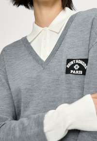 Persoon die een grijze V-hals trui draagt over een wit overhemd met knopen, met een patch "Saint Honore Paris" op de borst van de trui.