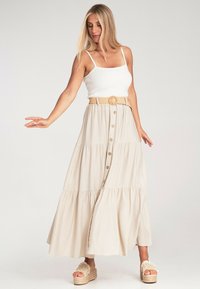 Beige Maxikleid mit gestuften Design und geknöpftem Vorderteil, kombiniert mit einem weißen, taillierten Tanktop und einem beige geflochtenen Gürtel, ergänzt durch Plateausandalen.