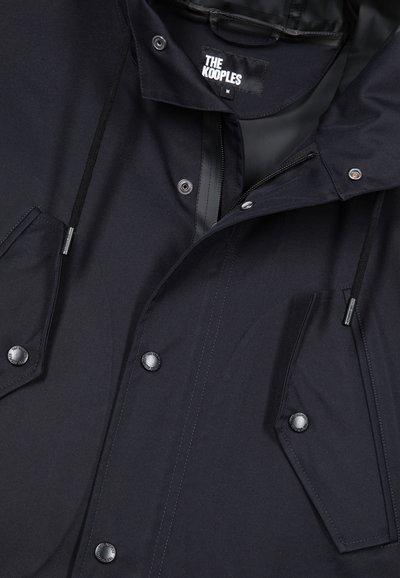 Veste légère noire avec boutons-pression, deux grandes poches avant, capuche réglable avec cordon de serrage et étiquette de marque à l'intérieur. Texture lisse.