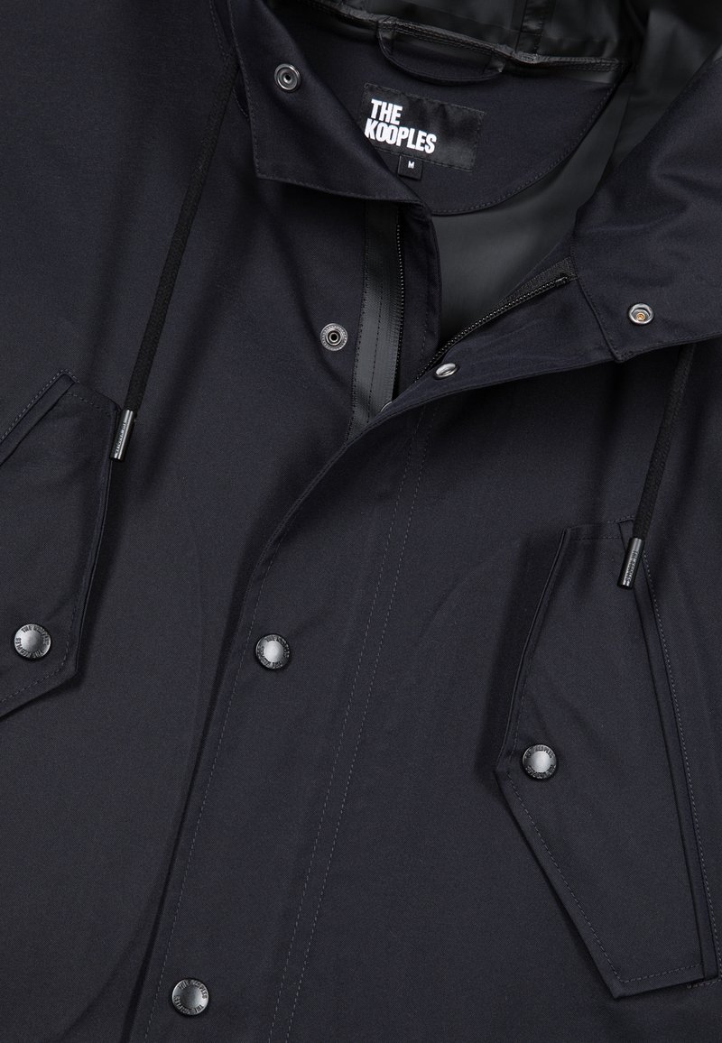 Chaqueta ligera negra con botones a presión, dos grandes bolsillos frontales, capucha ajustable con cordón y etiqueta interior de marca. Textura suave.