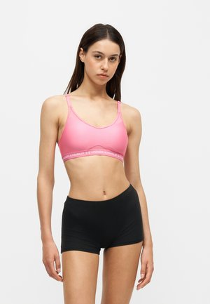 Frau trägt einen pinkfarbenen Sport-BH und schwarze Sportshorts und steht mit neutralem Gesichtsausdruck vor einem schlichten Hintergrund.
