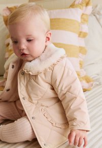 Manteau matelassé rose avec un col en fausse fourrure et des accents à volants, doté de boutons-pression argentés, porté avec des collants rose clair. Oreiller rayé en arrière-plan.