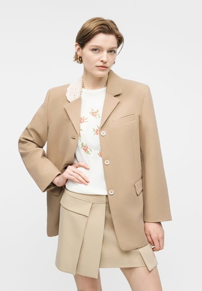 Νεαρή γυναίκα που φοράει μπεζ σακάκι, λευκό τοπ με λουλούδια και μπεζ πλισέ φούστα, ποζάροντας με το ένα χέρι στη μέση της μπροστά από ένα απλό φόντο.